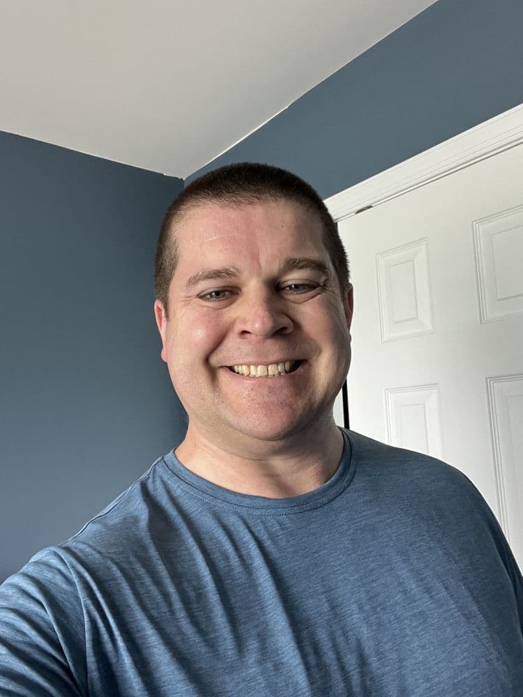 A smiling man in a blue athletic shirt stands in front of a white door with a blue wall in the background, suggesting a personal training or fitness environment associated with Tim Carrigan Personal Training LLC in Allentown, PA.
