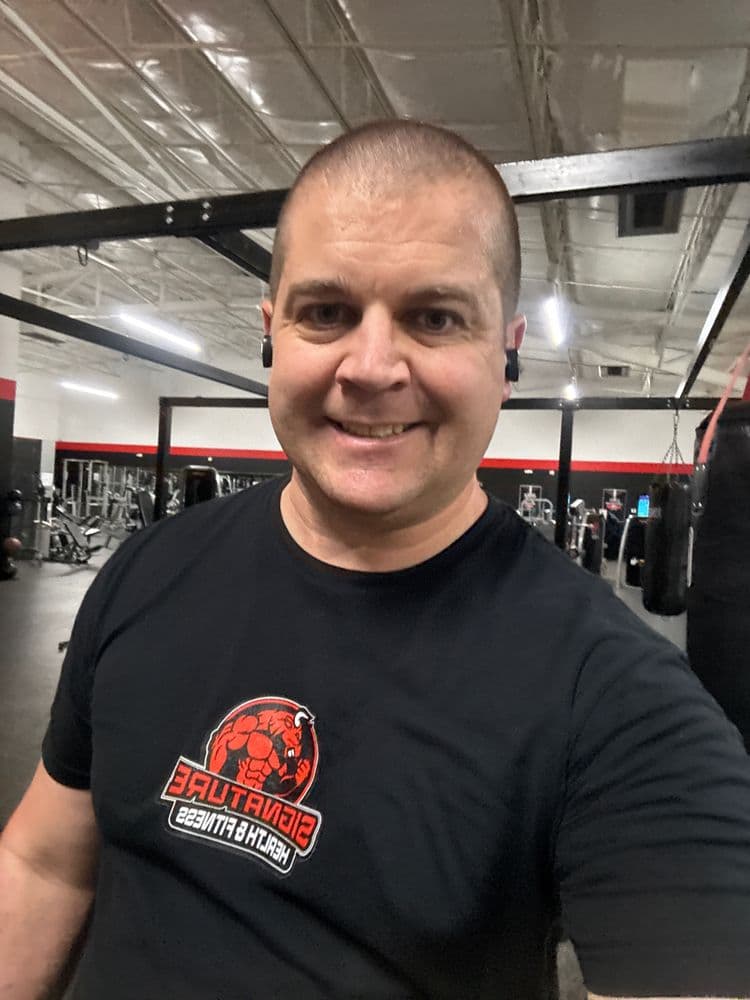 A smiling man wearing a black T-shirt with the "Signature Healthcare Fitness" logo, standing inside a gym with exercise equipment in the background.
